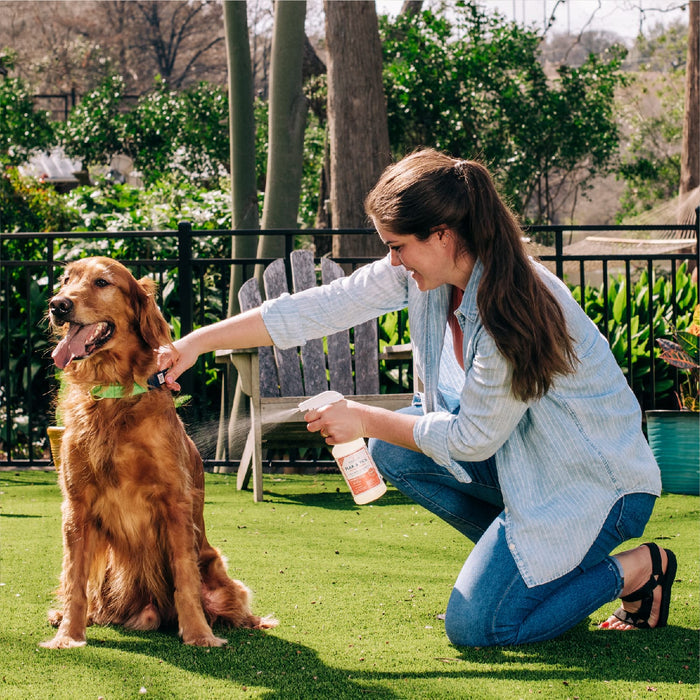 A woman kneels on grass, smiling as she sprays Wondercide 128 oz. Peppermint Flea & Tick Spray—safe for pets—onto a golden retriever in a green collar. Lush greenery and a wooden chair appear in the background.