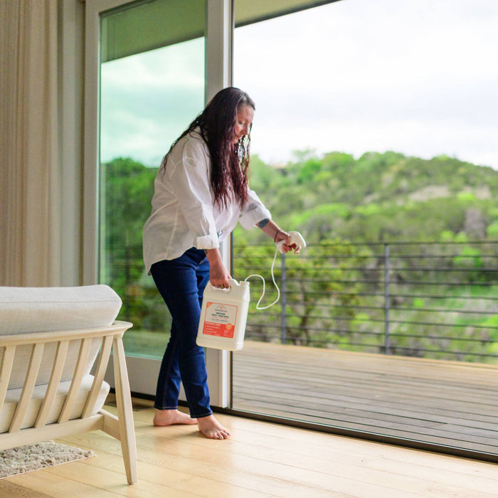 A woman in a white shirt and jeans uses Wondercide's Home Treatment Kit | Flea & Tick Spray from a large container on the floor near a glass door, with a scenic green home and yard visible in the background.