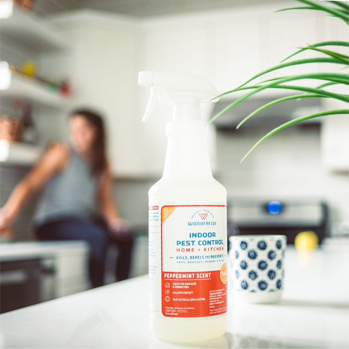 A 128 oz. bottle of Wondercide Peppermint Indoor Pest Control sits on a white kitchen counter beside green plant leaves, a patterned mug, and a blurred person—safe for pets and effective for indoor pest protection.