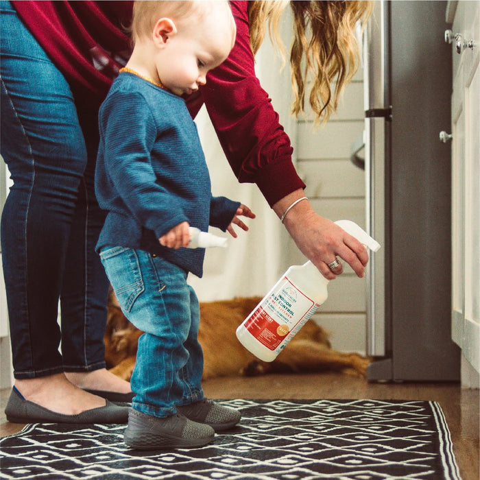 A toddler stands on a black and white rug with a spray bottle as an adult uses Wondercide 128 oz. Peppermint Indoor Pest Control; a brown dog relaxes by the cabinets, showing it’s safe for use around pets.