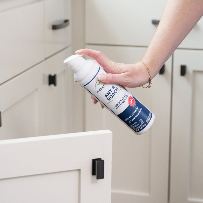 A person uses Wondercide (2 Pack) Ant & Roach for Home + Kitchen, a plant-powered spray that kills ants and roaches in a white kitchen cabinet. The blue and white can is safe around pets; cabinets have black handles, and the hand wears a gold bracelet.