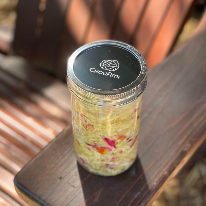 ChouAmi stainless steel flat panel sealing a Le Parfait jar of fermenting sauerkraut with rose petals on a wooden bench outdoors.