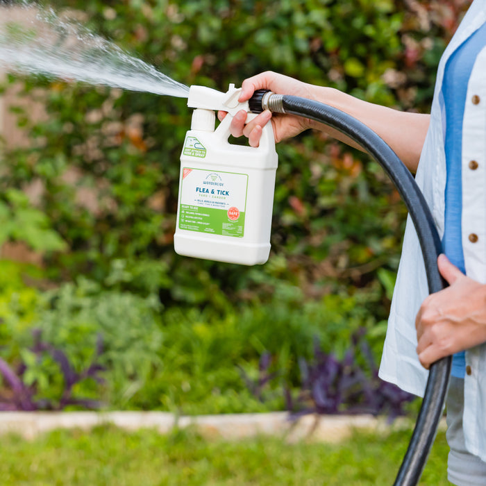 A person uses the Wondercide Complete Control Flea & Tick Kit, spraying plant-powered yard protection from a white bottle outdoors with green grass and plants visible in the background.