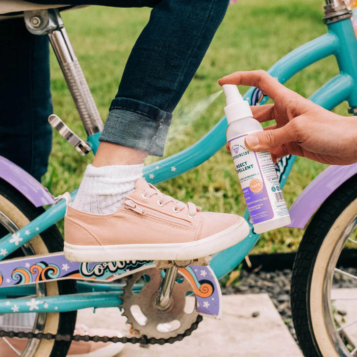A person applies Wondercide Insect Repellent for Family to a child's ankle as the child, in pink sneakers and rolled-up jeans, sits on a blue and purple bike outdoors, helping repel ticks and mosquitoes for safe playtime near the grass.