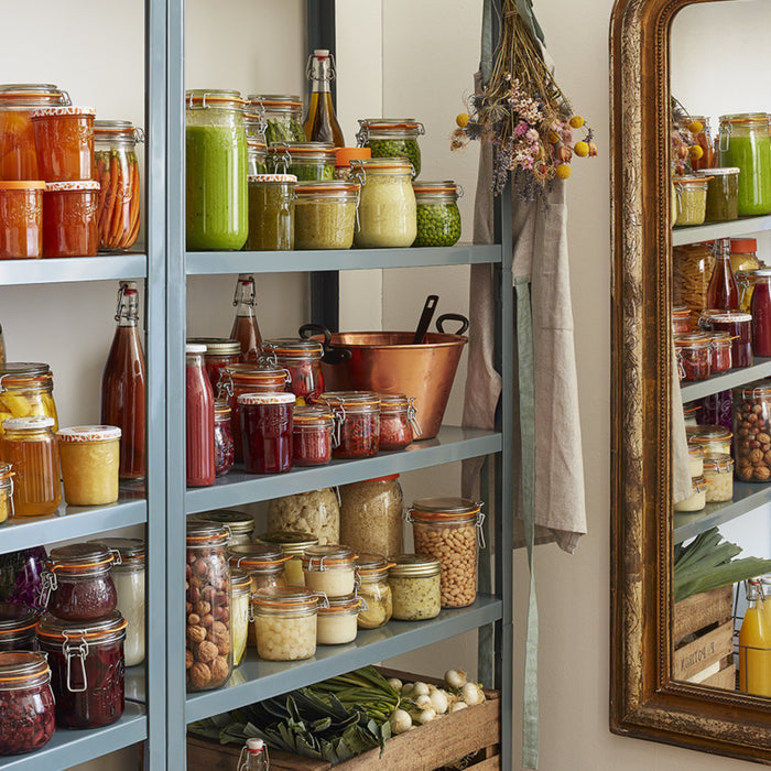 Pantry shelves stocked with Le Parfait jars and bottles of preserves and juices, with a copper bowl, vintage gold mirror, and dried flowers.