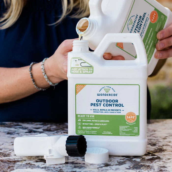 A person pours liquid into a white Wondercide Mosquito Yard Spray Refill Starter Kit container on a marble surface, with a hose spray attachment nearby. The label notes its power to kill, repel, and prevent outdoor pests.