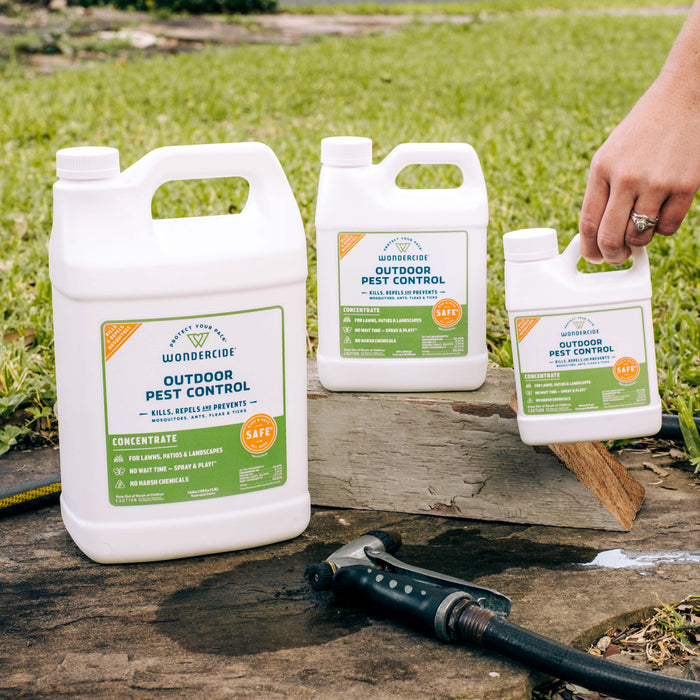 Three Wondercide Outdoor Pest Control Concentrate for Yard + Garden containers are on a wooden plank outside with grass in the background. A hand holds the smallest, and a garden hose nozzle—perfect for applying this mosquito repellent—is nearby.