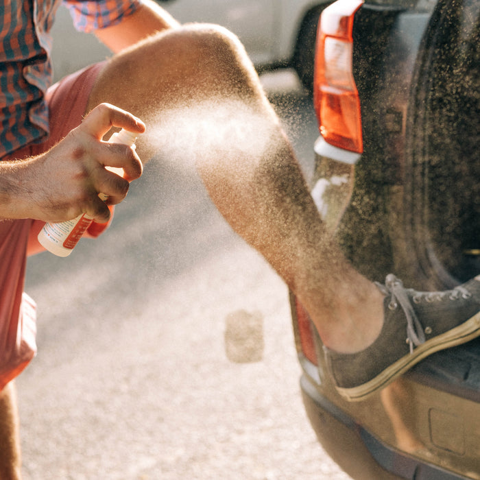 A person in shorts and sneakers sprays Wondercide Peppermint Insect Repellent for Family on their leg, enjoying its fresh peppermint scent while resting a foot on a car bumper in bright sunlight.