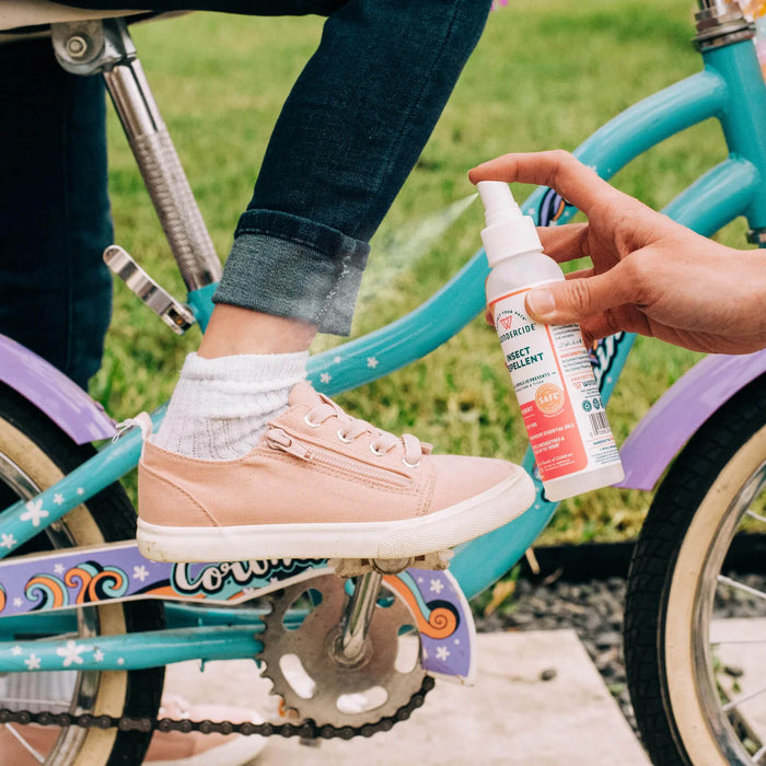 An adult sprays Wondercide Peppermint Insect Repellent for Family on a child’s ankle as the child, in pink sneakers and rolled-up jeans, sits on a blue and purple bicycle.