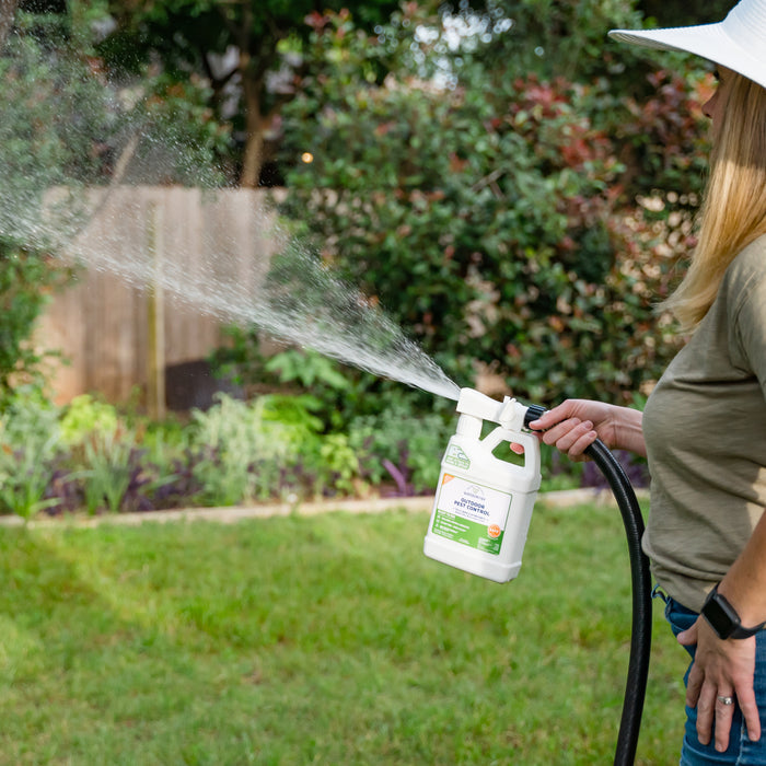 A woman in a wide-brimmed hat uses Wondercide Ready-to-Use Outdoor Mosquito Spray with a hose-attached bottle, treating backyard plants by the wooden fence to help kill mosquitoes and other pests.