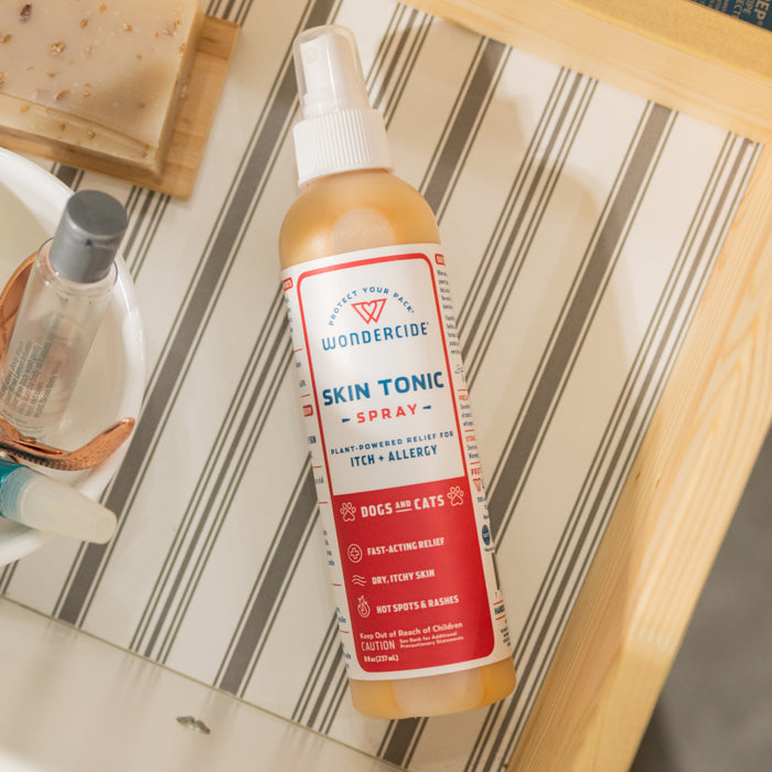 A bottle of Wondercide Skin Tonic Itch Spray sits on a striped surface next to a soap bar, small bowl, and other bathroom items.