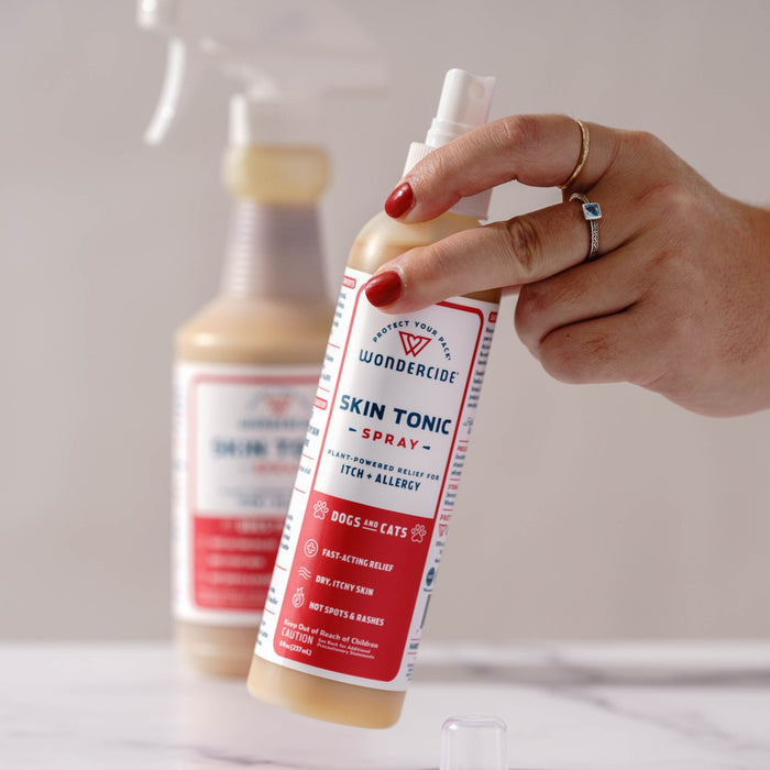 A hand with red nail polish holds Wondercide Skin Tonic Itch Spray for pets, formulated to soothe itching, with another Wondercide spray bottle blurred in the background on a light surface.