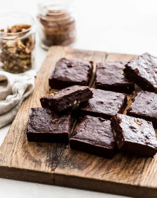Chocolate brownies cut into squares on a wood board with chopped walnuts and a jar of cocoa.