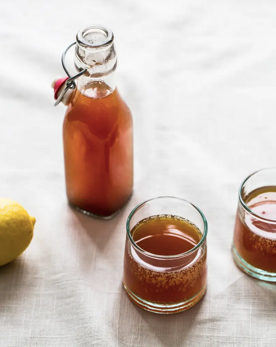 Swing-top bottle and two glasses of amber kombucha beside a fresh lemon on a linen tablecloth.